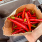 Hand holding a paper bag of red peppers with a background of tomatoes and a wooden surface.